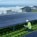 Wide photo showing agrivoltaic solar panels above crop rows with a researcher inspecting plants, modular battery storage containers mid-scene, and in the distance a coastline with wave-energy buoys and a small community solar array near suburban homes under bright overcast light.