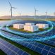 Wide, slightly elevated view of a solar farm with wind turbines and white battery storage containers, linked by subtle luminous arcs symbolizing AI optimization at golden hour, with distant transmission lines and mountains.