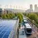 Elevated wide shot of a city block with rooftop solar arrays, electric buses charging, bioswales and bike lanes along the street, and distant wind turbines at golden hour.