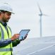 Renewable energy professional in high-visibility gear reviewing a tablet beside solar panels with wind turbines in the distance under an overcast sky.