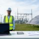 “Analyst in a hard hat holding a tablet in front of wind turbines and solar panels, representing renewable energy asset management.”