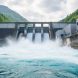 “A hydropower dam with a river flowing over the spillway, showing water mist and a powerhouse in a green valley under an overcast sky.”