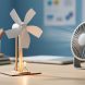 Close-up of a student STEM windmill model with paper-cup rotor blades spinning near a small fan, while a small LED glows to demonstrate wind energy conversion.