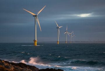 Offshore wind turbines on the U.S. East Coast at dusk, viewed from the shoreline with choppy water in the foreground.