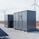 Grid-scale battery energy storage cabinets at a solar farm, with solar panels and wind turbines softly visible in the distance.