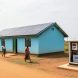 Rural off-grid renewable energy installation with solar panels on a clinic roof and a small wind turbine nearby, with community members walking in the background.