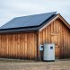 Remote cabin with a rooftop solar panel array and nearby off-grid power equipment in a rural landscape under overcast light.
