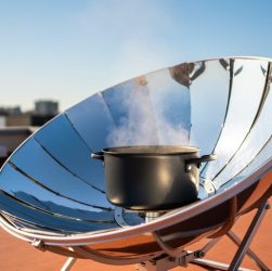 Solar cooker with a reflective parabolic dish focusing sunlight onto a dark pot on a rooftop, with steam rising in bright daylight.
