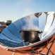 Solar cooker with a reflective parabolic dish focusing sunlight onto a dark pot on a rooftop, with steam rising in bright daylight.