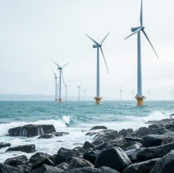Offshore wind turbines generating clean power in the ocean near a rocky coastline under an overcast sky.