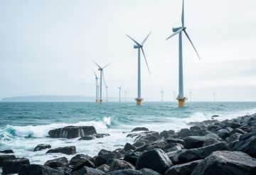 Offshore wind turbines generating clean power in the ocean near a rocky coastline under an overcast sky.